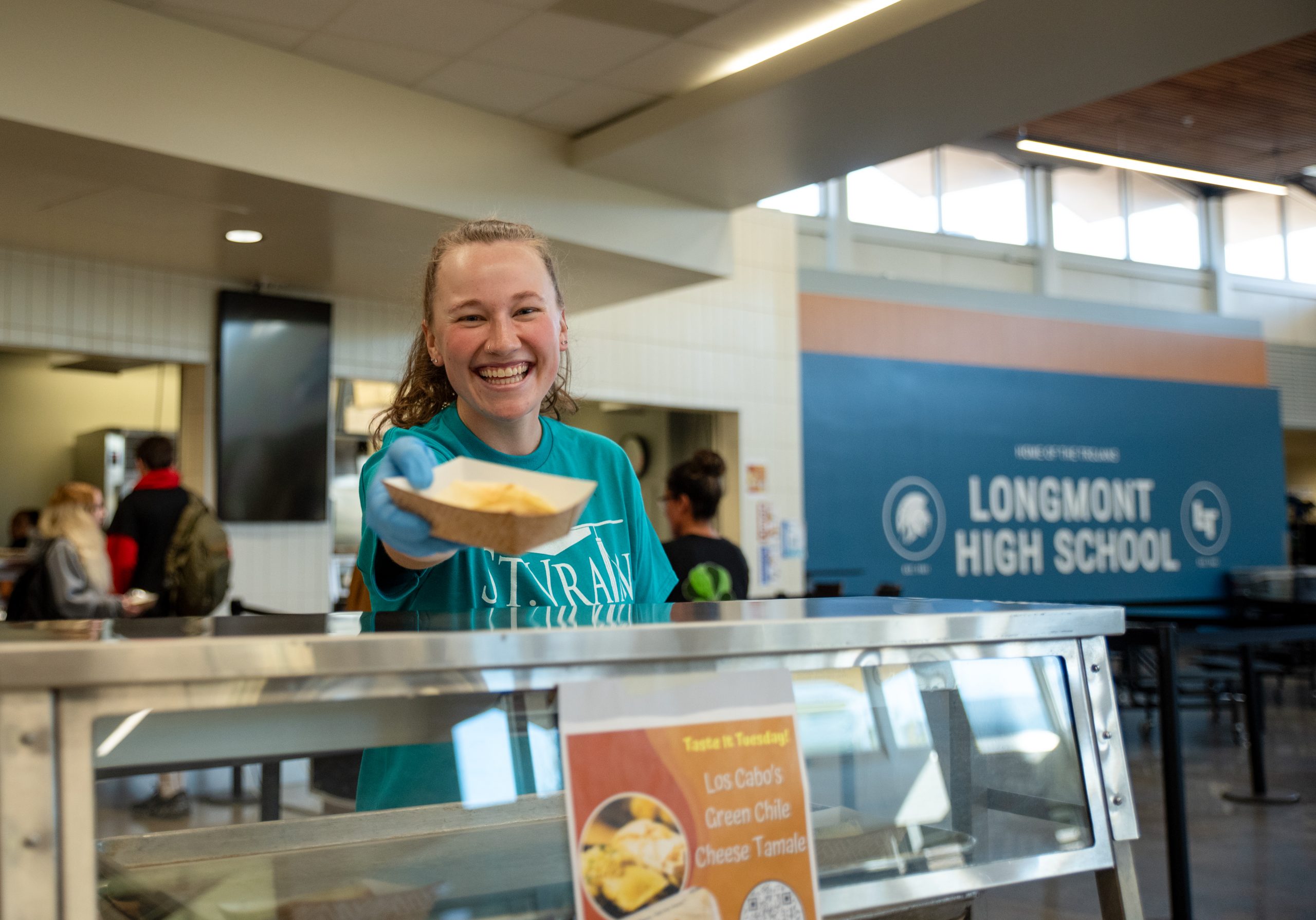 Un estudiante sonriente en una cafetería sostiene una bandeja con comida, con un letrero del colegio al fondo.