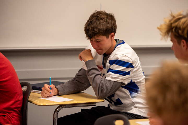 Un estudiante con una camiseta de fútbol está sentado en un pupitre, escribiendo en un papel con expresión concentrada en un aula.