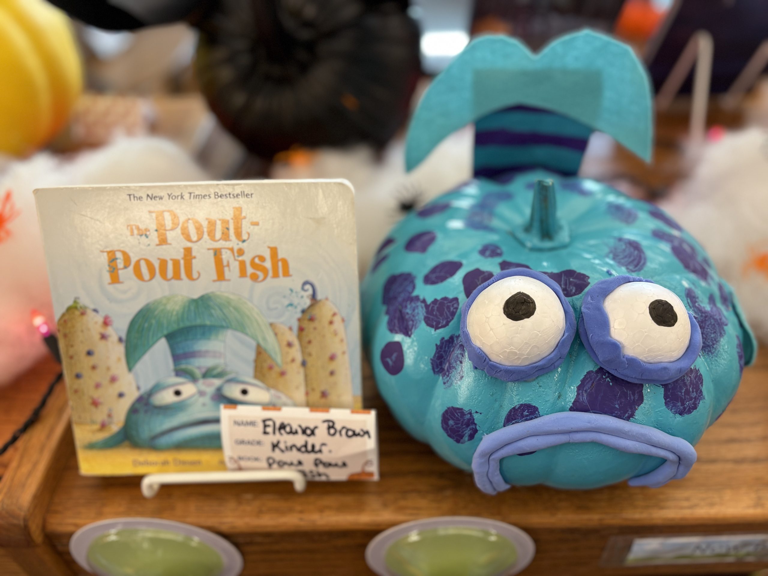 Una calabaza azul decorada como el pez Pout-Pout junto al libro «El pez Pout-Pout», con una etiqueta con su nombre.