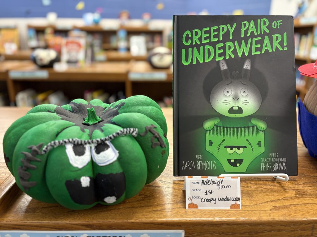 Una calabaza verde con cara junto al libro «¡Un par de calzoncillos espeluznantes!» de Aaron Reynolds, sobre una mesa de la biblioteca.