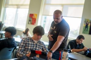 Un profesor ayuda a un alumno con una tableta en el aula, mientras que el resto de alumnos trabajan en sus pupitres.