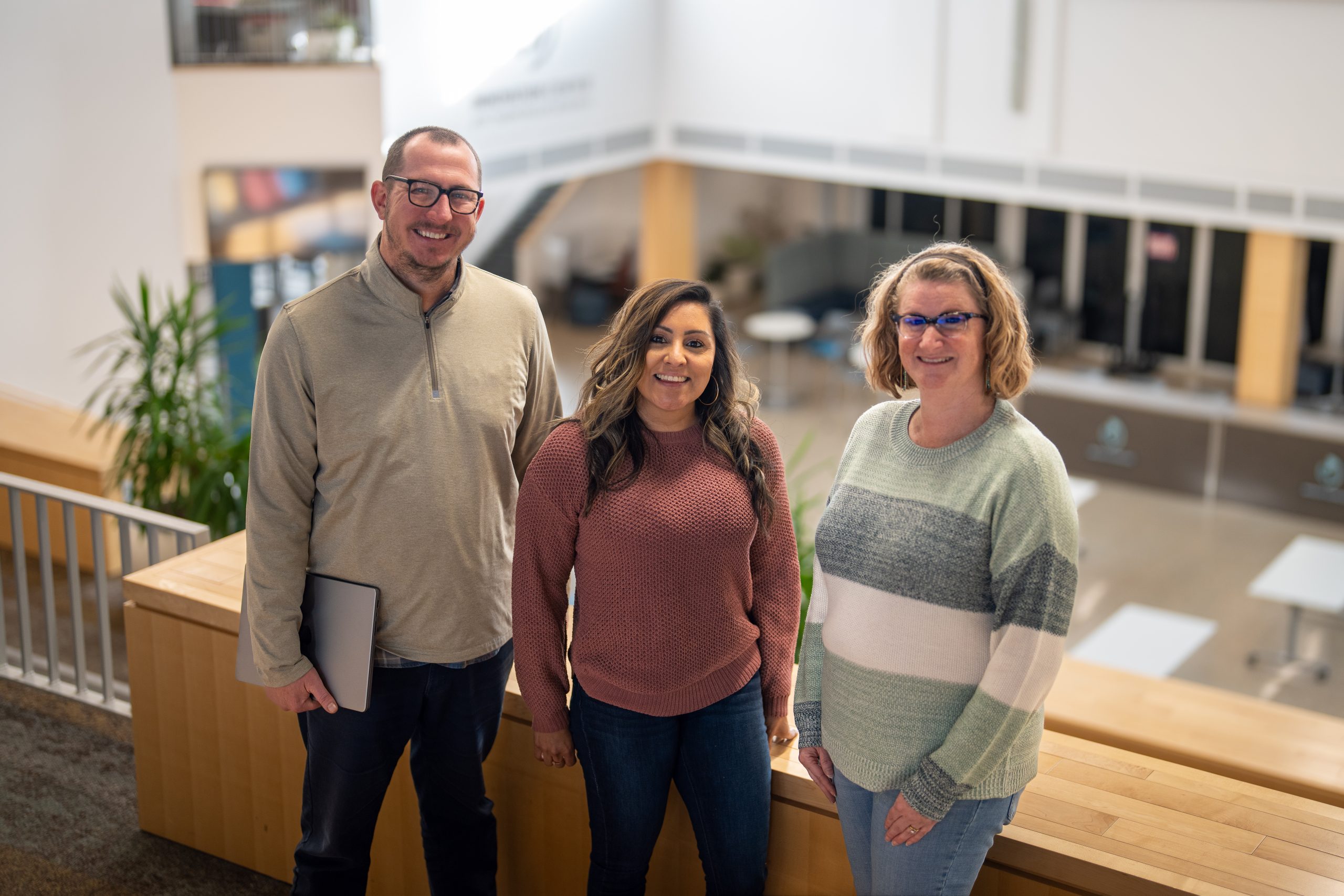 Three people stand together in a modern indoor space, smiling. They wear casual sweaters and jeans, with greenery in the background.