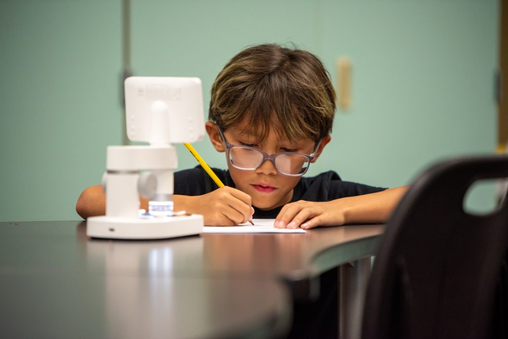 Un niño pequeño con gafas escribe con un lápiz en una mesa, concentrado en su trabajo, con un microscopio a su lado.