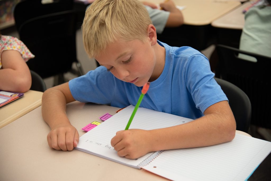 Un niño con una camiseta azul escribe en un cuaderno en el pupitre de un aula, concentrado en su trabajo.