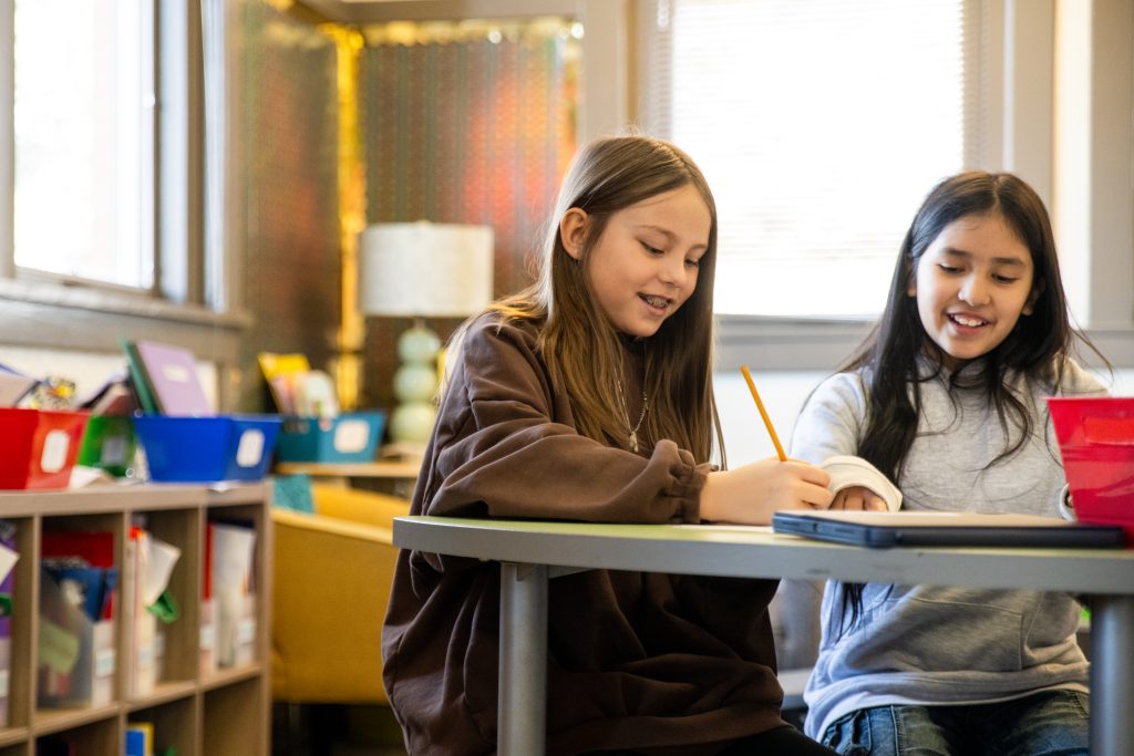 Dos niñas sonrientes están sentadas en una mesa en un aula, escribiendo y compartiendo ideas. Cerca de ellas hay unas cajas de colores.