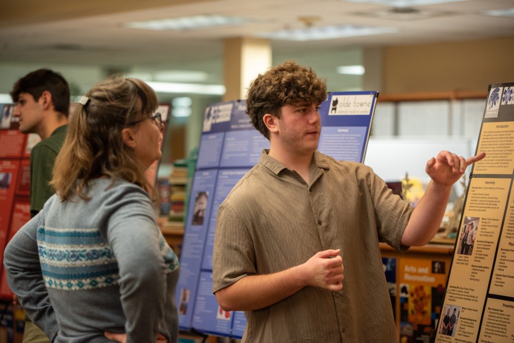 A student explains a poster to a woman at a presentation event, with informational displays in the background.
