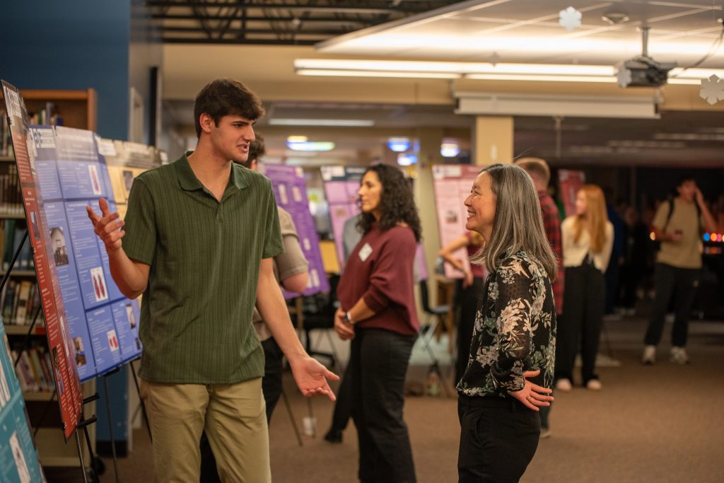 Two people engage in conversation at a poster presentation event, surrounded by attendees and informational displays.