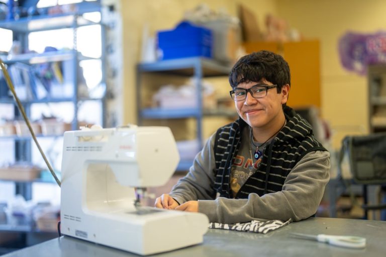 Una alumna sonríe sentada ante una máquina de coser, rodeada de materiales de manualidades en un aula.