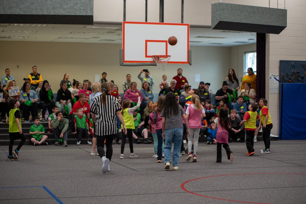 Un partido de baloncesto juvenil en pleno desarrollo, con jugadores vestidos con camisetas de colores y un público animando al fondo.