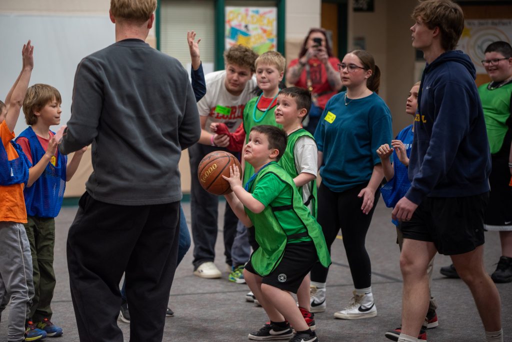 Un grupo de niños en un gimnasio; un niño vestido de verde se dispone a lanzar una pelota de baloncesto mientras los demás observan emocionados.