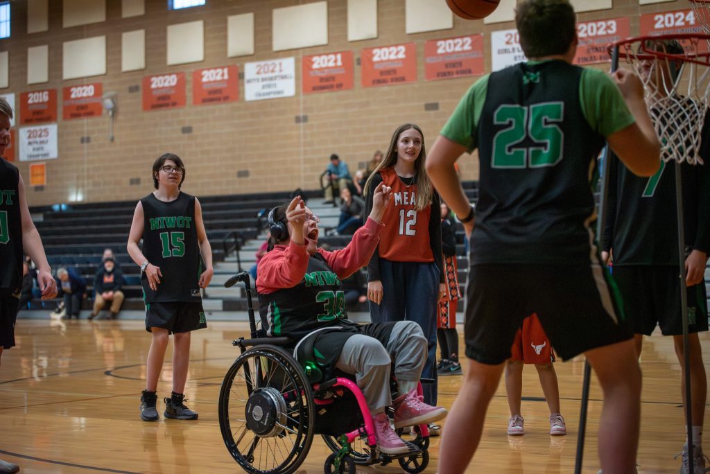 Una niña en silla de ruedas juega al baloncesto, rodeada de sus compañeras de equipo, que llevan camisetas, mientras lanzan el balón hacia la canasta.