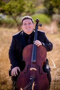 Un músico sonriente, vestido con un traje negro, está sentado al aire libre, con un violonchelo en las manos, rodeado de hierba alta y vegetación.