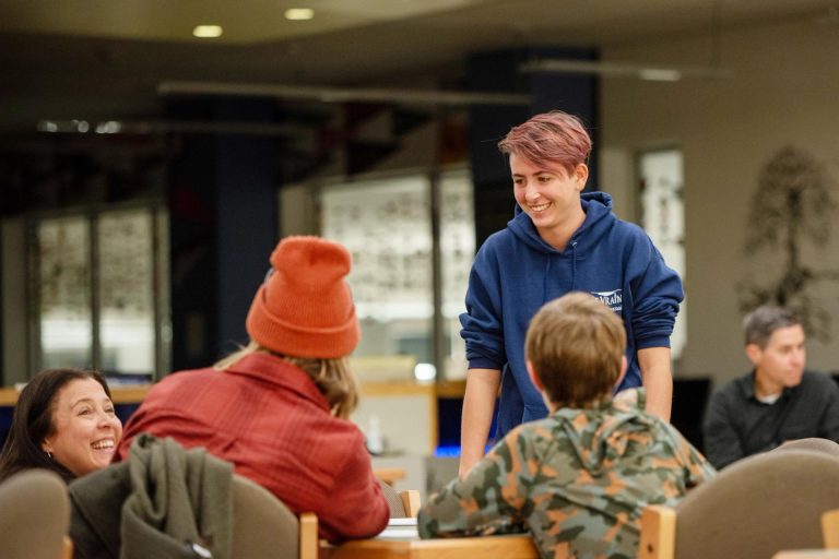 Un grupo de personas conversando en una mesa, con una persona sonriente vestida con una sudadera azul con capucha de pie cerca de ellos.
