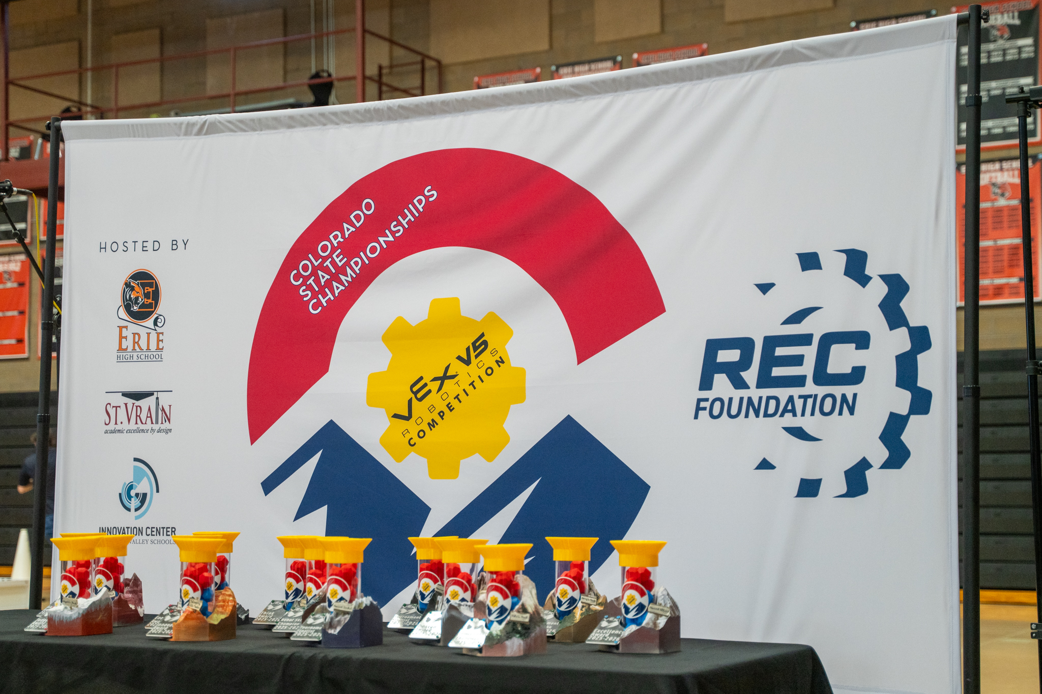 Cartel del Campeonato Estatal de Colorado con los logotipos de VEX Robotics y trofeos de colores sobre una mesa.
