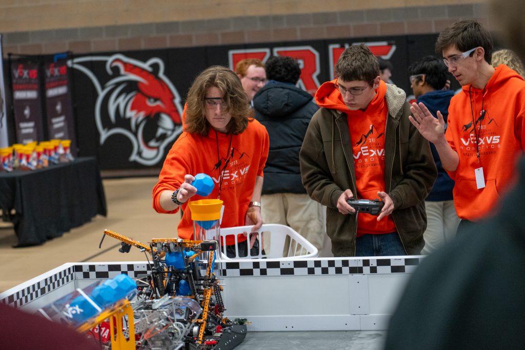 Tres estudiantes con sudaderas naranjas con capucha manipulan el montaje de una competición de robótica, centrados en el control de un robot.