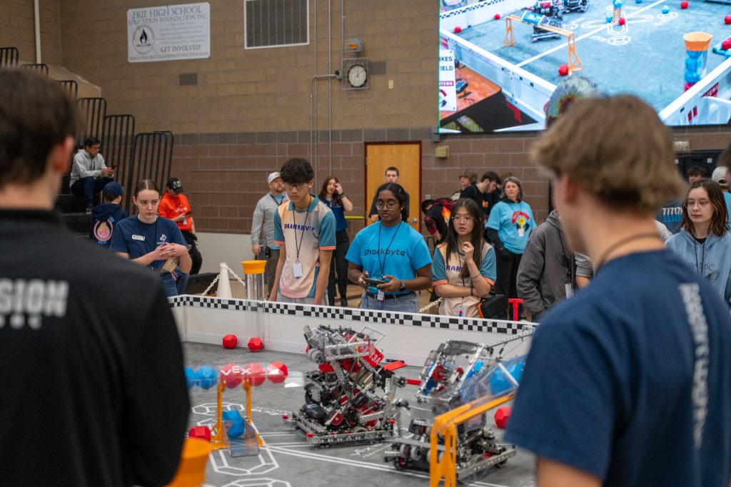 Los alumnos observan una competición de robótica, con robots en una pista y una gran pantalla en la que se retransmite el evento.