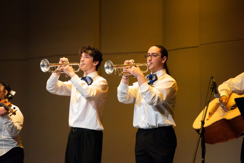 Two musicians play trumpets on stage, wearing white shirts with blue accents, while a violinist is partially visible.