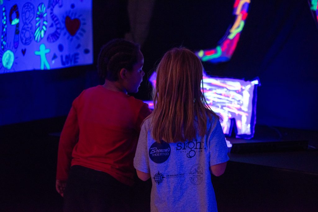 Dos niños observan unas coloridas obras de arte iluminadas con luz negra, rodeadas de decoraciones llamativas.