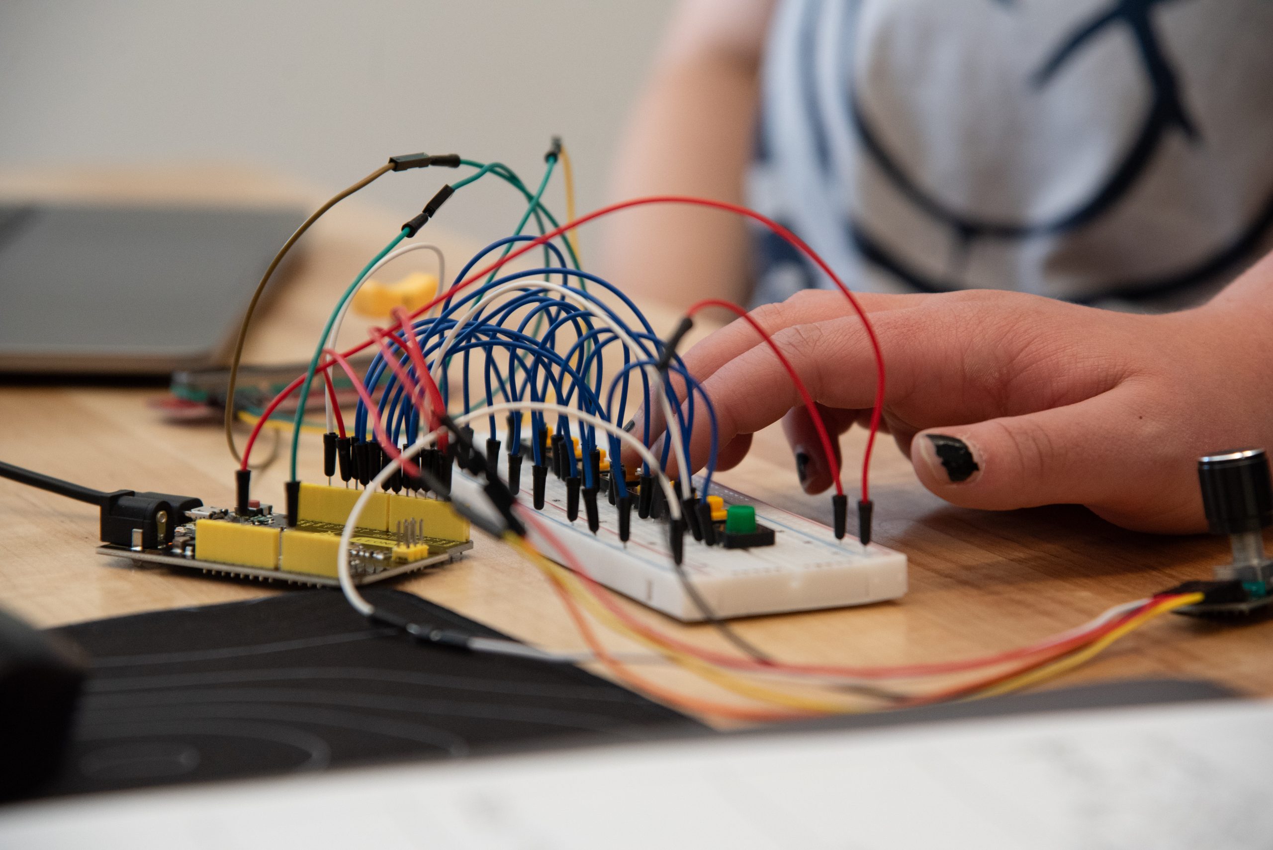 Una mano conecta los cables de una placa de pruebas con un microcontrolador y diversos componentes sobre una mesa de madera, tocando un prototipo de instrumento musical.