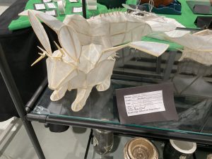 A white origami-style sculpture resembling an animal, displayed on a glass shelf with an artist's information card.