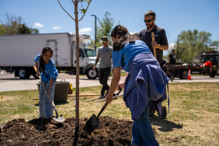 Dos niños están plantando un árbol mientras los adultos les ayudan en una iniciativa de jardinería comunitaria en un día soleado.