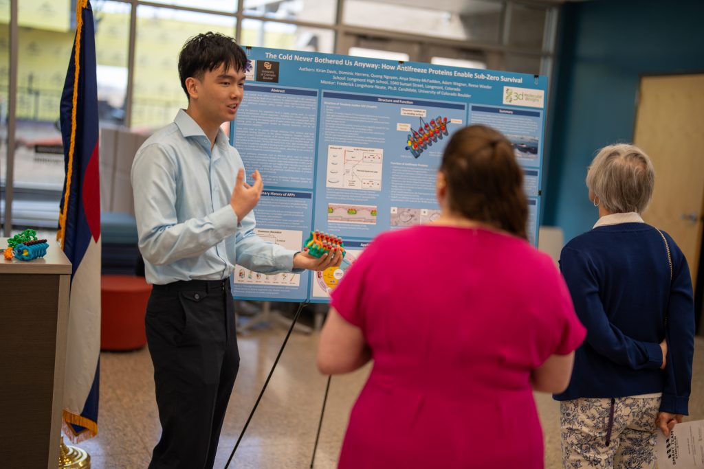 Un joven presentador explica su investigación sobre las proteínas anticongelantes a dos asistentes en un evento científico.
