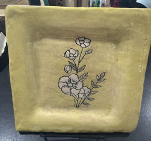 A square yellow plate featuring a floral design with outlined flowers and leaves, displayed on a stand.