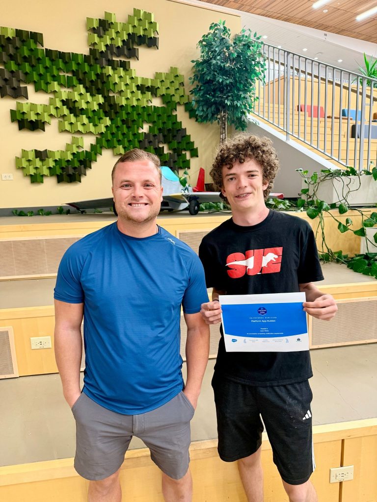 Two young men stand smiling, one holding a certificate, with a green wall design and plants in the background.