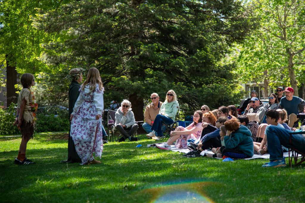 Los niños actúan en el escenario de un parque mientras un público compuesto por familias los observa desde el césped en un entorno soleado.