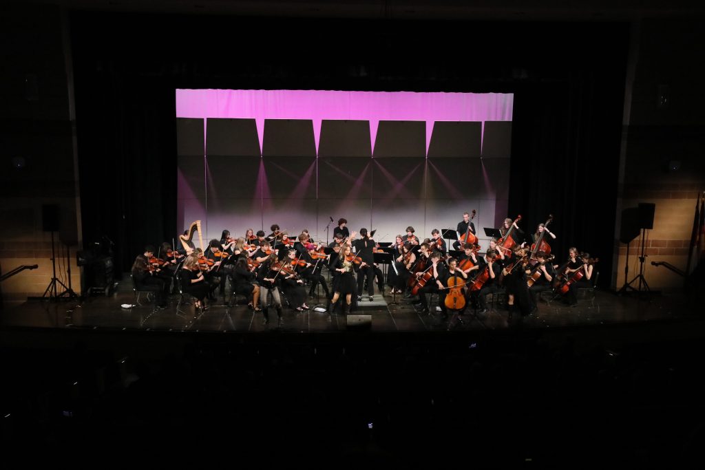 La orquesta Silver Creek High actúa en el escenario, con instrumentos de cuerda y músicos vestidos de negro bajo una iluminación colorida.