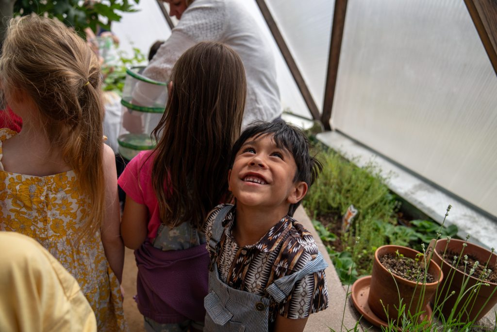 A smiling child looks up while surrounded by other children in a greenhouse filled with plants.