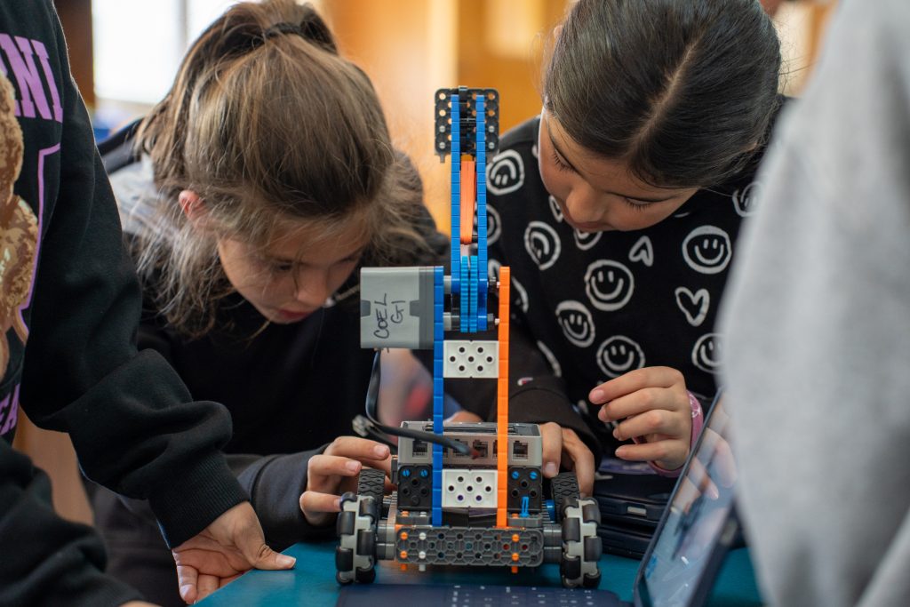 Dos niños se concentran intensamente en construir un robot con bloques de colores sobre una mesa, rodeados de sus compañeros de clase.