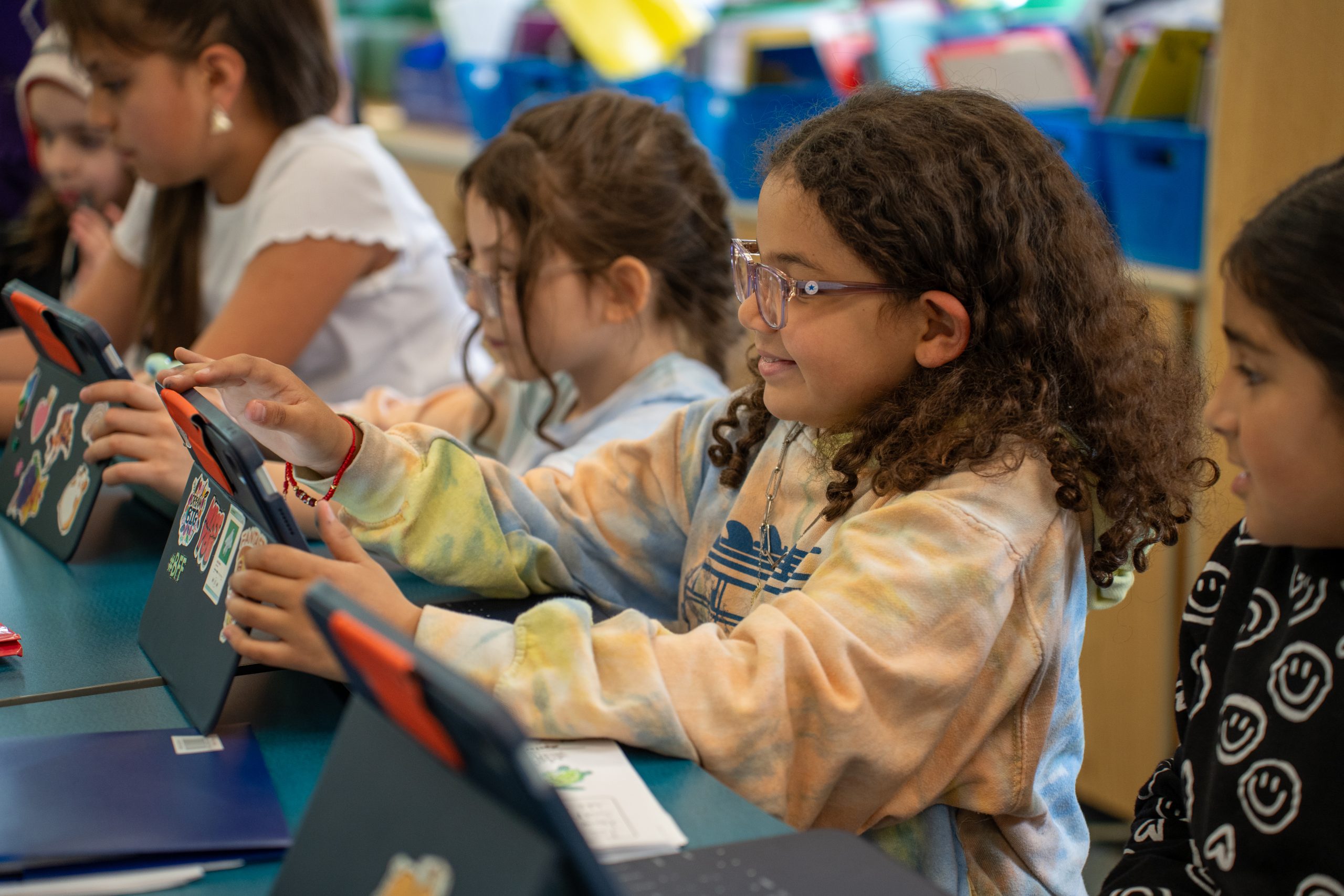 Students in a classroom are engaged with tablets, focused on their work, with colorful stickers on the devices.