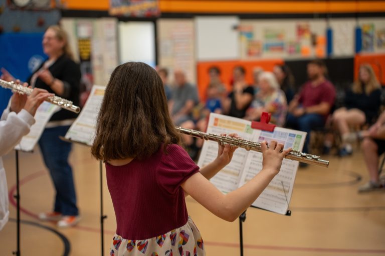 A young girl plays the flute in a gym, with music sheets in front of her and an audience in the background.
