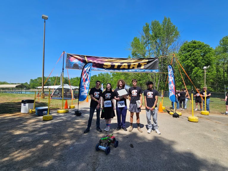 Un equipo de seis personas se encuentra en la línea de salida de una competición de rovers, sosteniendo una maqueta de rover bajo un cielo azul y despejado.