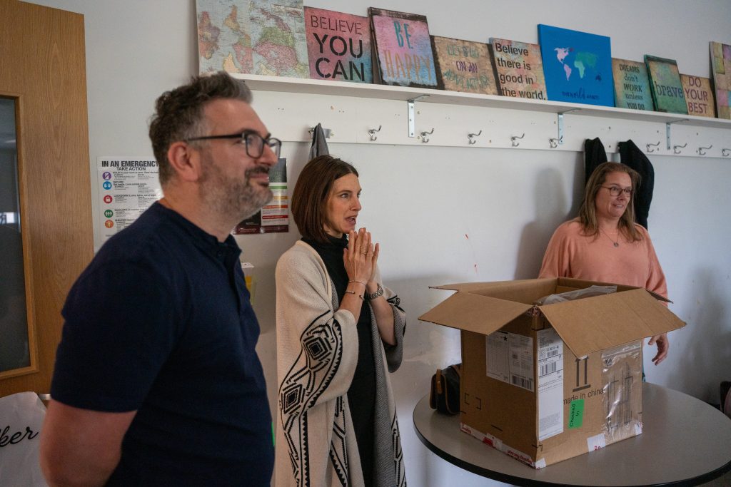 Three people stand in a room with a cardboard box on a table, surrounded by motivational wall art.