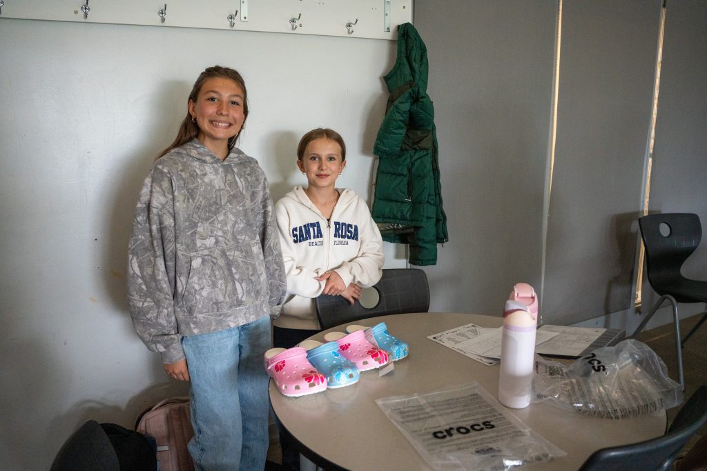 Two smiling girls stand by a table with colorful Crocs, a water bottle, and papers in a classroom setting.