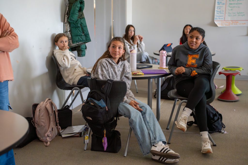 Students sitting in a classroom, engaged in conversation, with backpacks and notebooks around them.