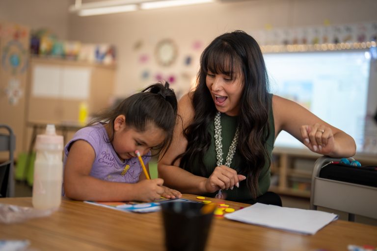 A teacher and a young girl engage in a creative activity at a table, both smiling and focused on their work.