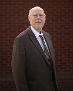 A man with glasses and a beard stands in a brown suit against a brick wall background, smiling confidently.
