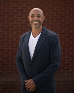 Smiling man in a black blazer over a white shirt stands against a red brick background.