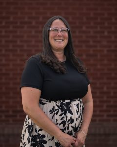 A smiling woman with long hair, wearing glasses, a black top, and a floral skirt, stands against a brick wall.