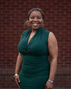 A smiling woman with braided hair wears a green dress, standing in front of a brick wall.