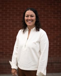 Smiling woman with curly hair in a white blouse stands in front of a brick wall, hands relaxed at her sides.