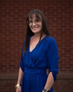 A woman in a blue dress smiles against a brick background, showcasing her glasses and long hair.
