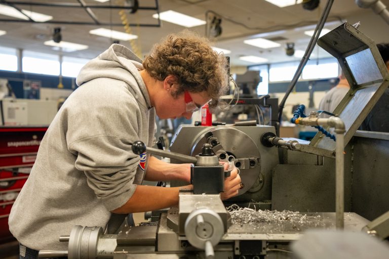A student works on a lathe machine in a workshop, focusing intently while wearing safety glasses. Shavings scatter around.