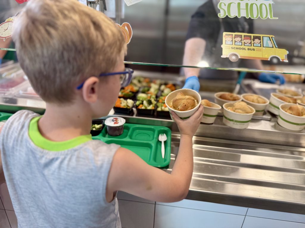 A child holds a bowl of food at a cafeteria while an adult serves food from behind a counter.