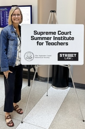 A woman stands beside a sign for the Supreme Court Summer Institute for Teachers, smiling in a casual outfit.