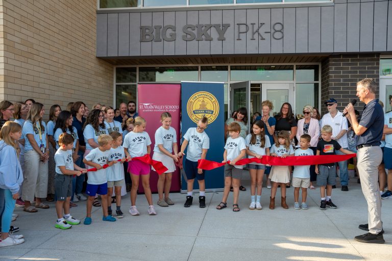A group of children and adults gather outside a school, preparing to cut a red ribbon for the opening ceremony.