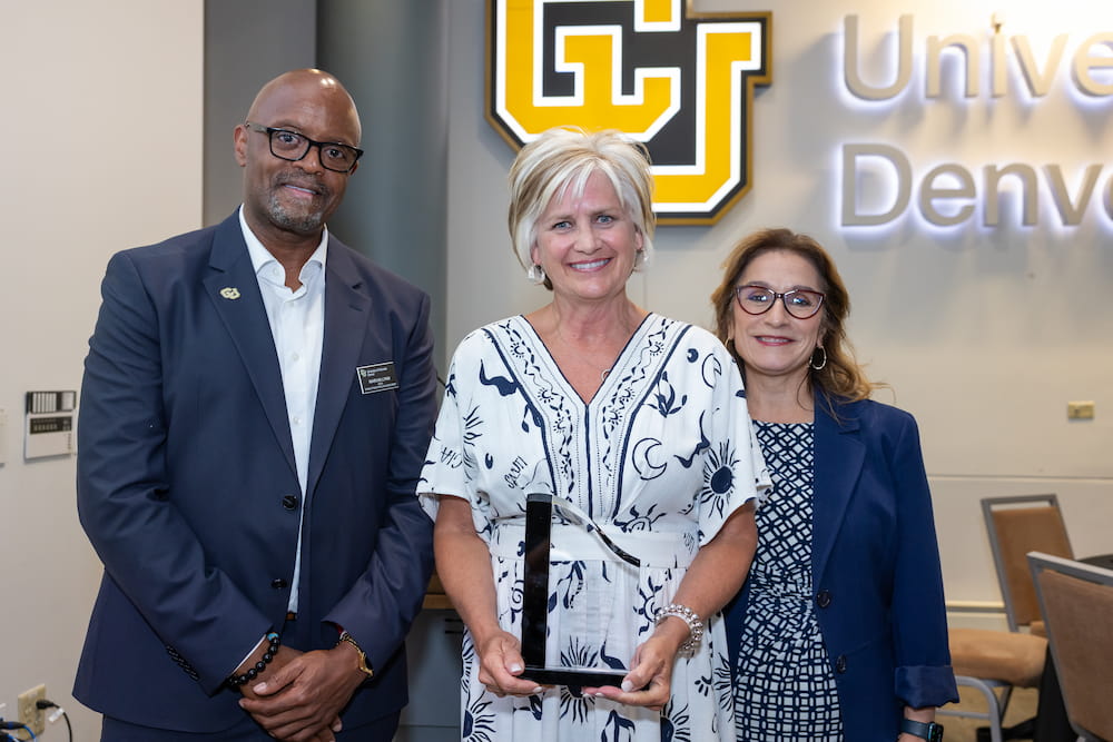 Three people pose together at an event, with one holding an award, in front of the University of Denver logo.